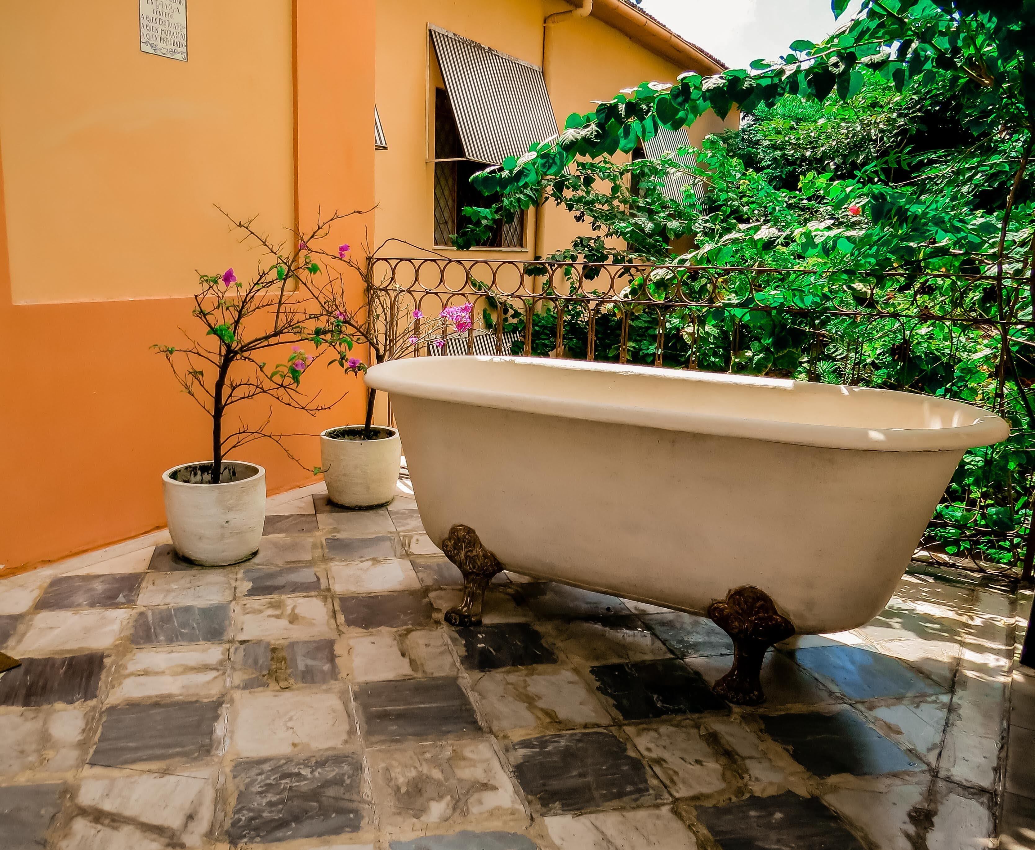 Una bañera antigua en un patio empedrado con plantas en macetas y una ventana con toldo a rayas.