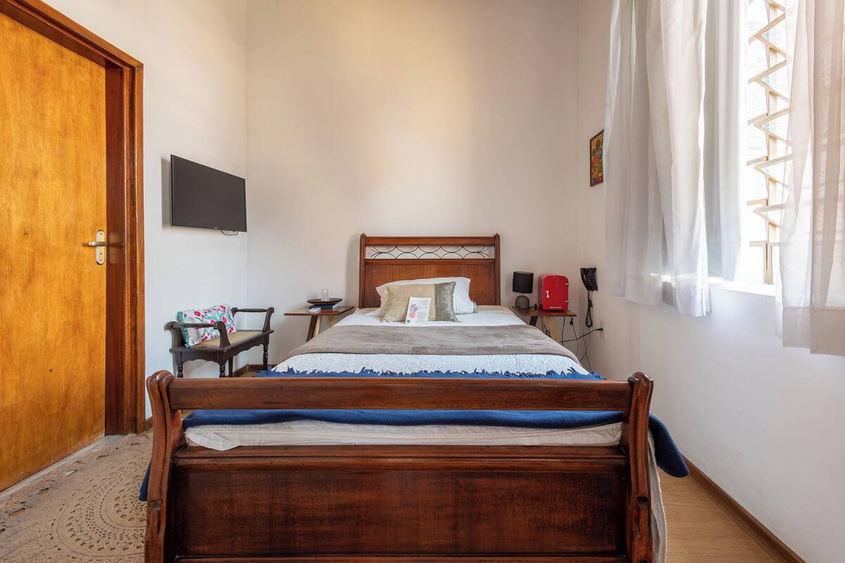 Une chambre  à coucher avec un lit en bois, un téléviseur fixé au mur, une table de chevet et une fenêtre avec des rideaux.