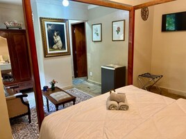 Quarto com cama de dossel, escrivaninha com cadeira, mesinha com um vaso e televisão na parede.