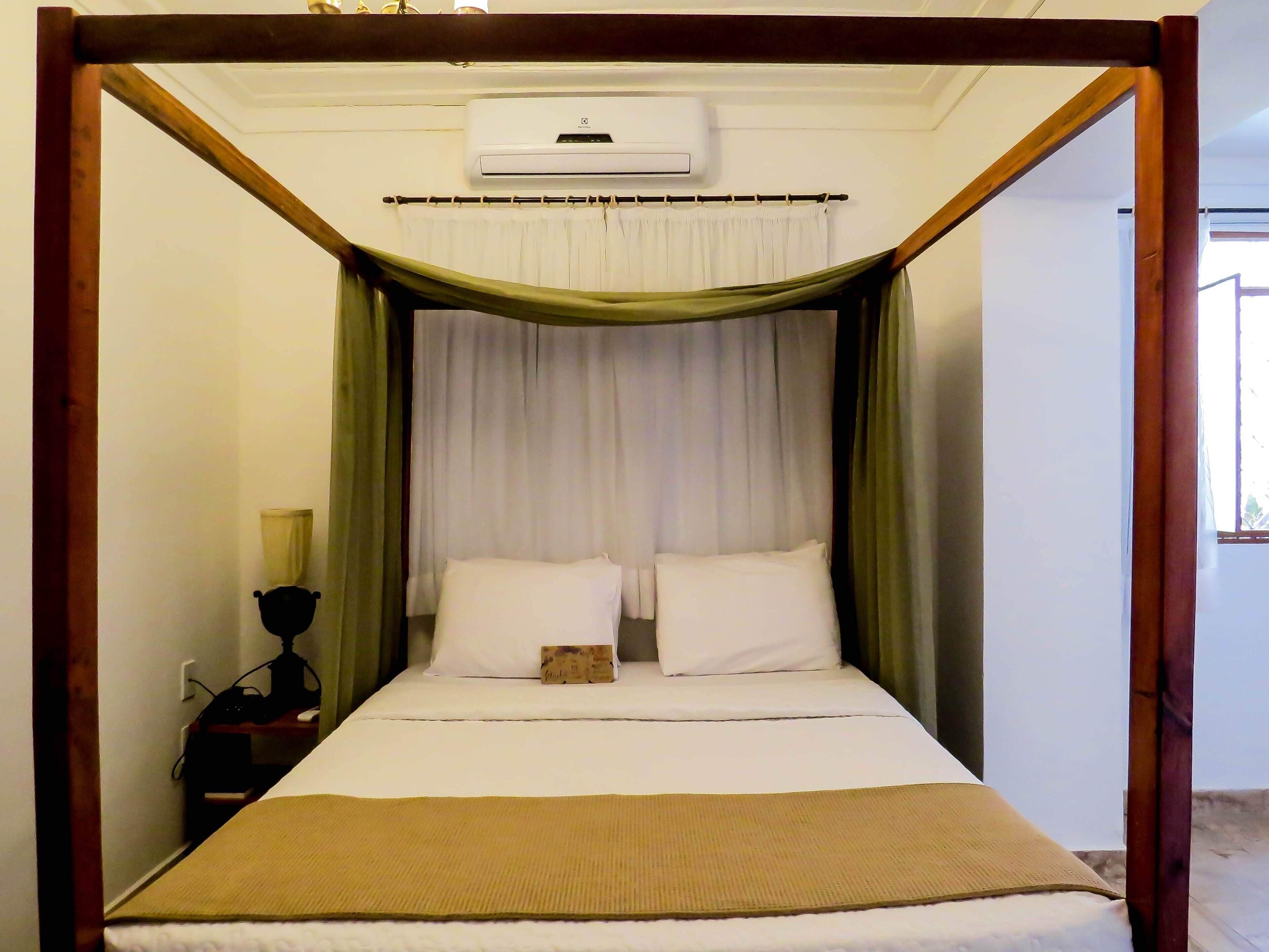 A four-poster bed with white linens and a green canopy, an air conditioning unit above, and a bedside table with a lamp.