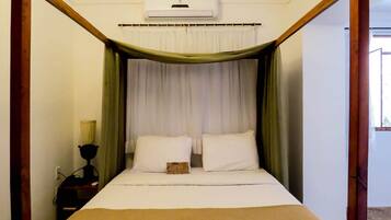 A four-poster bed with white linens and a green canopy, an air conditioning unit above, and a bedside table with a lamp.