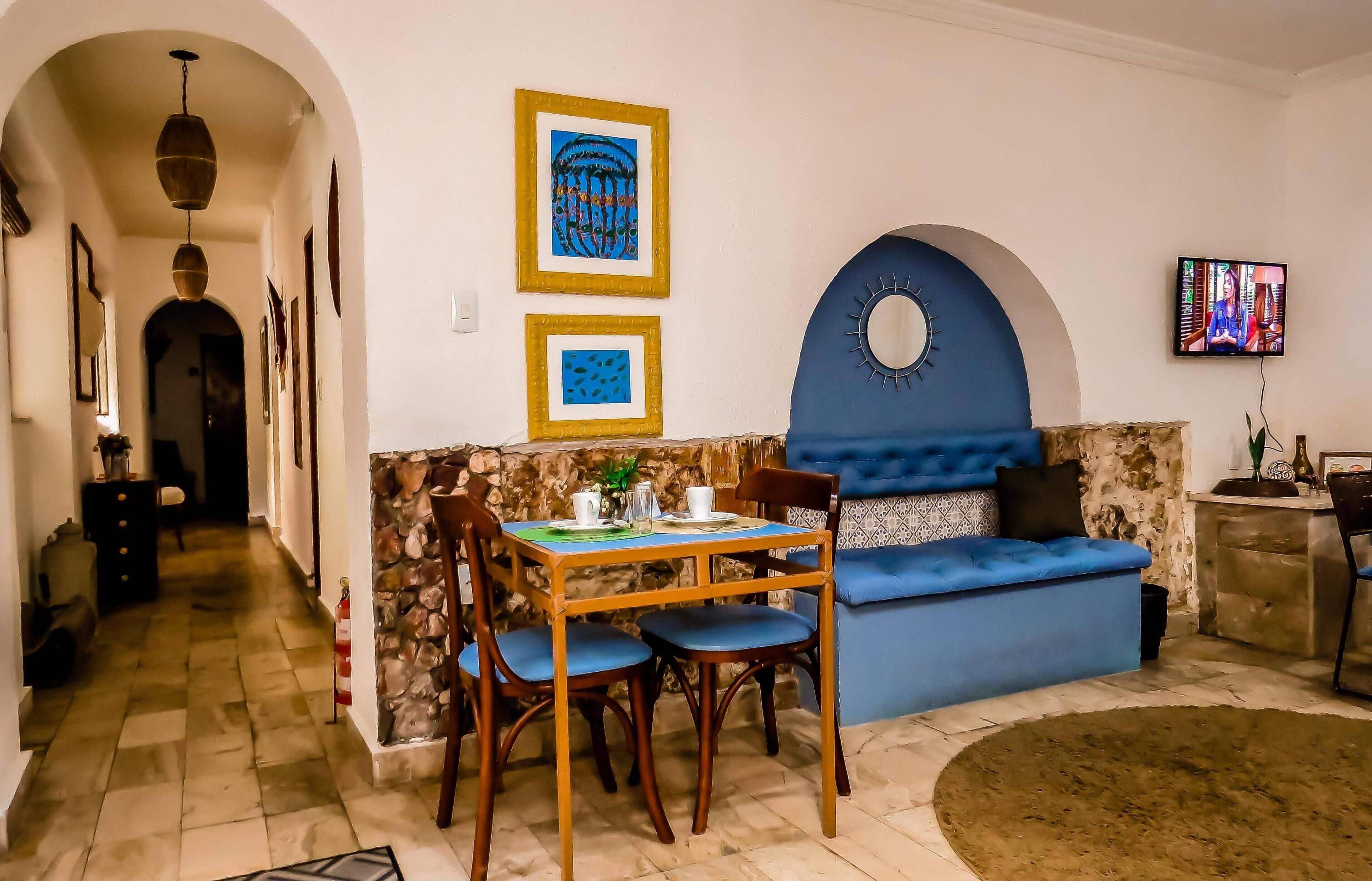 A cozy living area with a blue sofa, a dining table with chairs, and a stone wall. There are framed pictures on the wall and a TV mounted above the sofa.