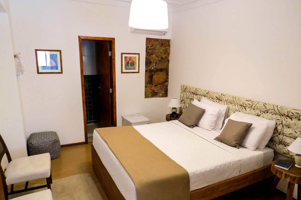 Une chambre à coucher bien rangée, avec un grand lit, une table de chevet avec une lampe, une chaise et une petite table sur laquelle se trouve un livre.