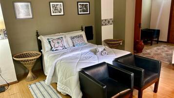 A bedroom with a bed, two chairs, a small table, a wall-mounted TV, and framed pictures.