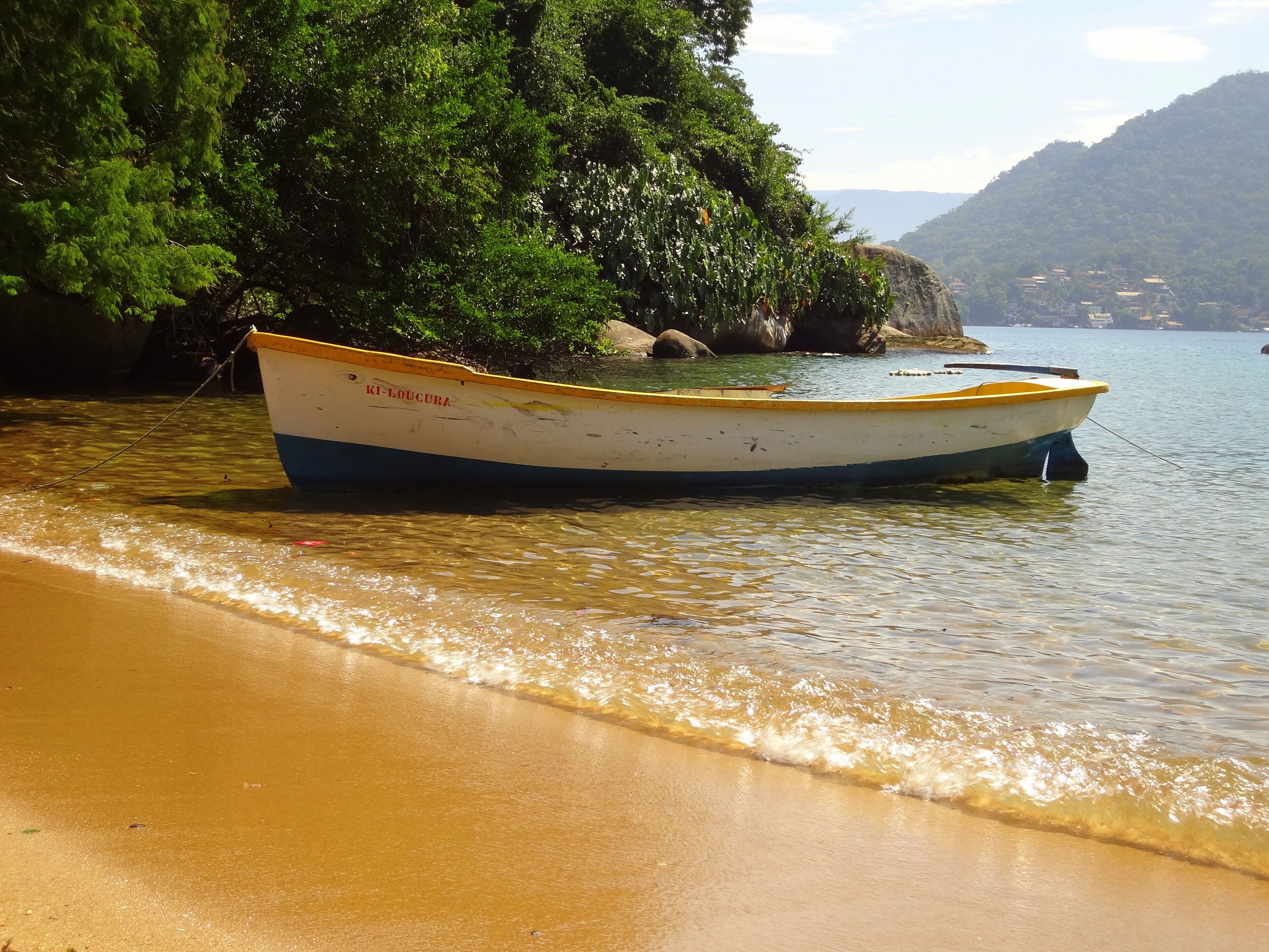 Un bateau nommé « KI-LOUCURA » était amarré sur une plage de sable fin, bordée d’une eau cristalline et dominée par un paysage montagneux.