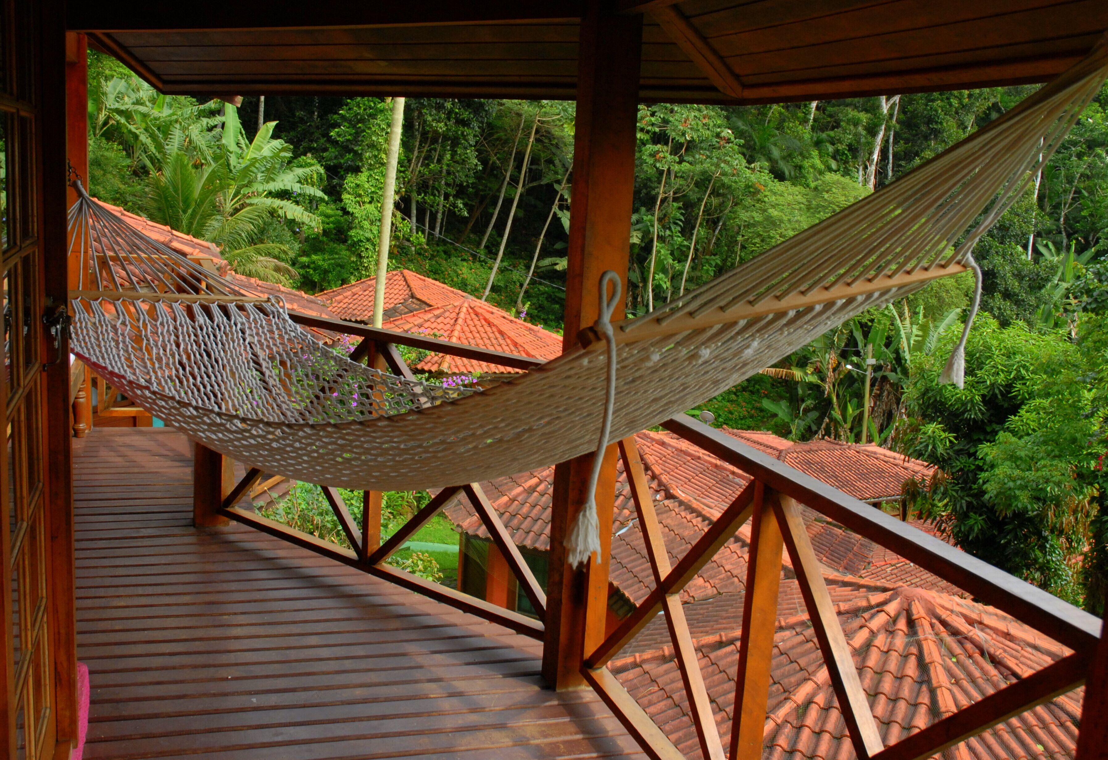 Un hamac sur une terrasse en bois donnant sur un jardin luxuriant, surmonté d’un toit recouvert de tuiles rouges.