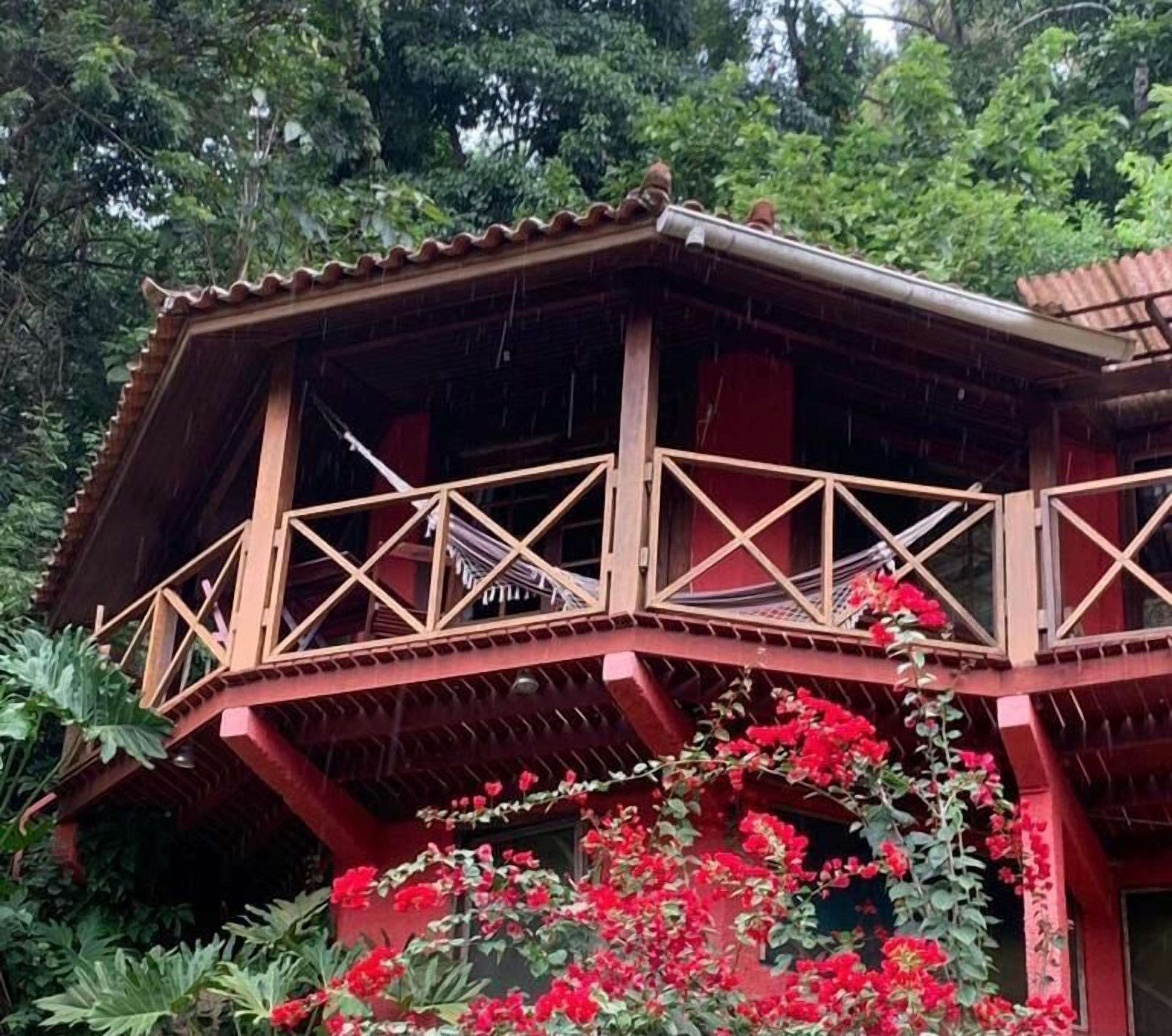 Una estructura de madera con balcón, rodeada de exuberante follaje y flores rojas.