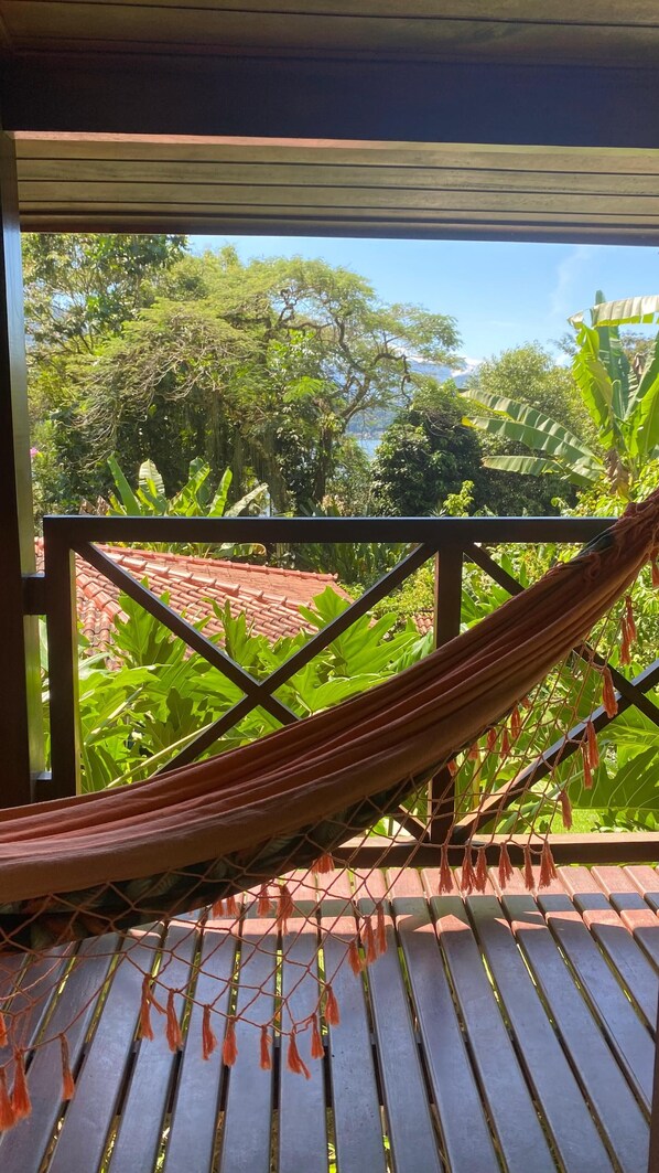 A hammock on a wooden deck with a view of lush greenery and a tiled roof in the background. - Pousada Vitorino (Angra dos Reis)