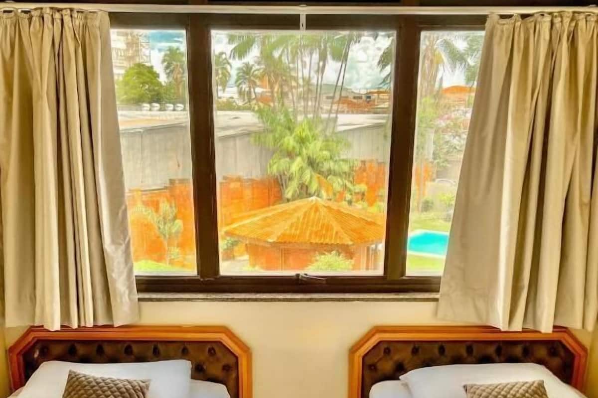 A room with two beds, a window with curtains, and a view of a palm tree and building outside.