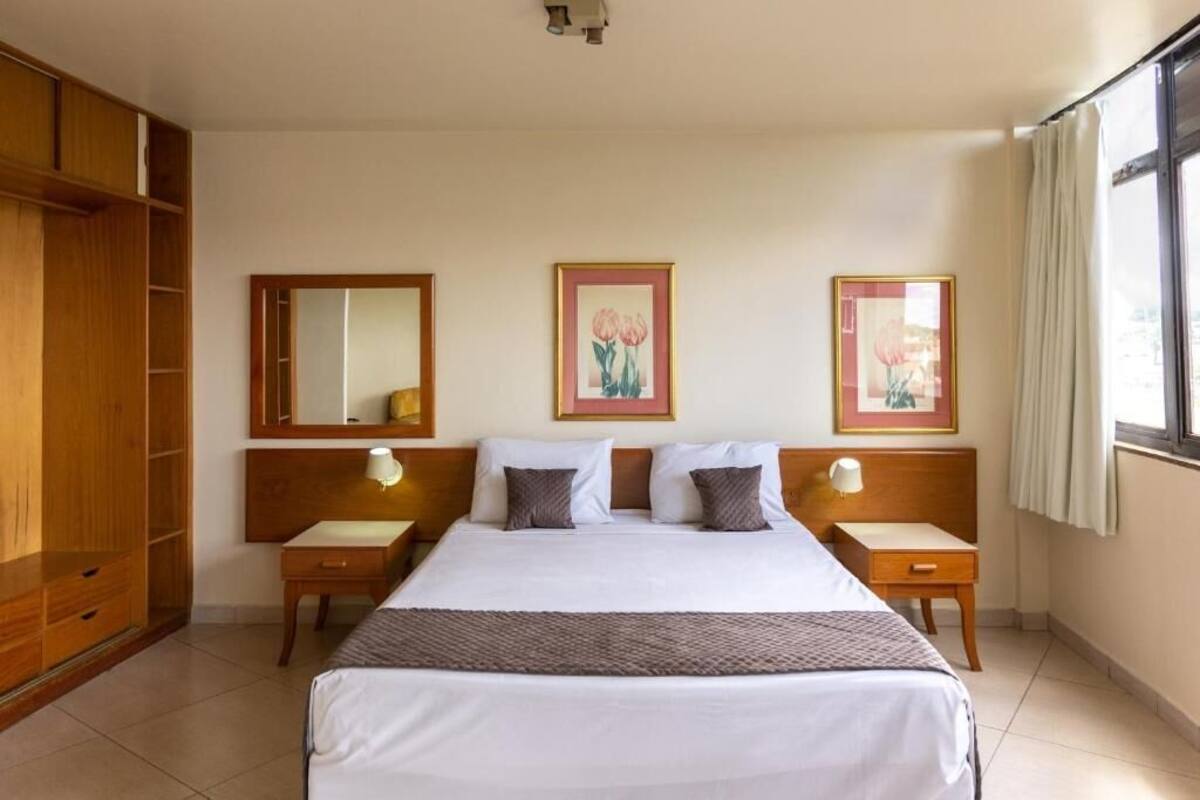 A hotel room with a large bed, wooden wardrobe, bedside tables with lamps, and framed floral artwork on the wall.