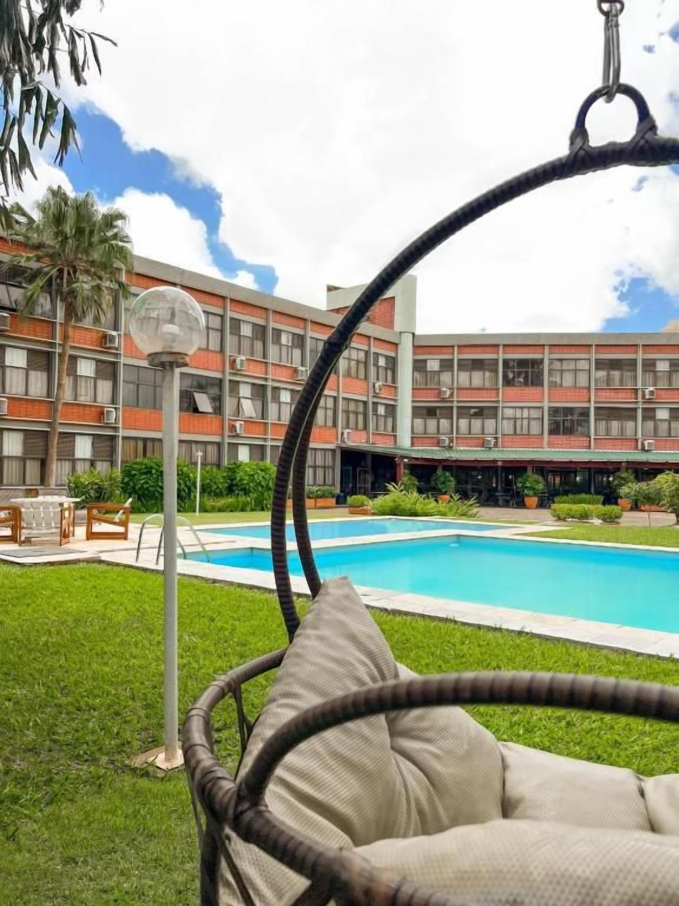 Un espace piscine agrémenté d’un fauteuil suspendu, un bâtiment doté de nombreux balcons et un ciel dégagé.