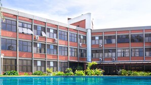 Il s’agit d’un bâtiment moderne à plusieurs étages, doté de grandes fenêtres et de climatiseurs. Devant le bâtiment se trouve une piscine avec une eau d’un bleu cristallin.