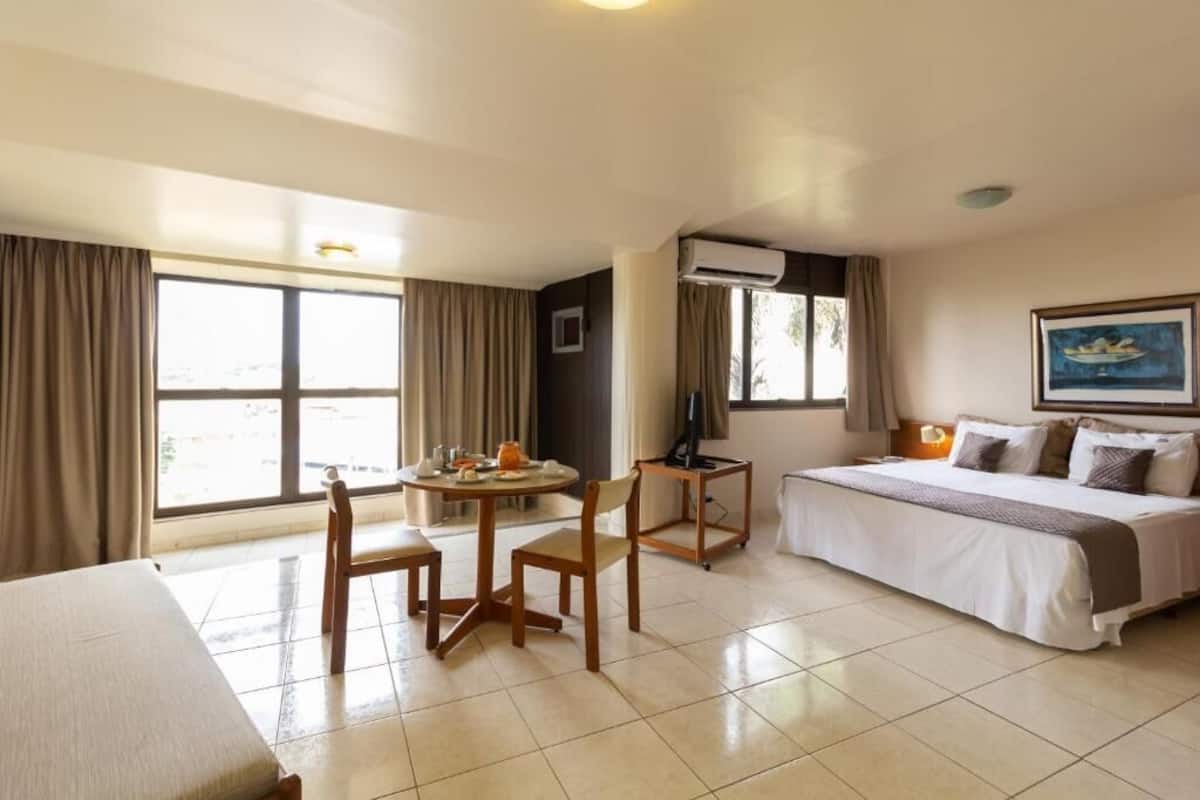 A hotel room with a bed, a desk, a chair, a table with a lamp, and a window with curtains.