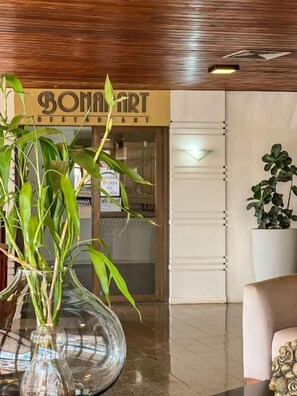 Eine Lobby mit einer Glaskaraffe grüner Pflanzen, einem Schild für 'BONAMART RESTAURANT' und einer großen Topfpflanze.