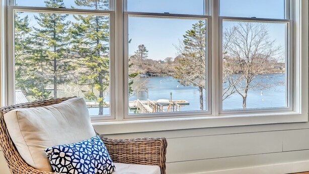 A wicker chair with a patterned pillow by a large window overlooking a lake with dock and trees.