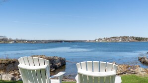Two white Adirondack chairs facing a calm body of water with a bridge in the distance and a shoreline with buildings.