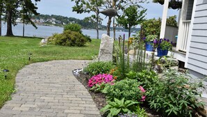 A paved pathway leading to a house with a garden featuring various plants and flowers.
