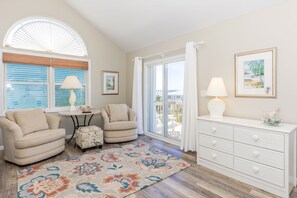 A room with a sofa, a small table with a lamp, a patterned rug, a white dresser, and a large window with blinds.