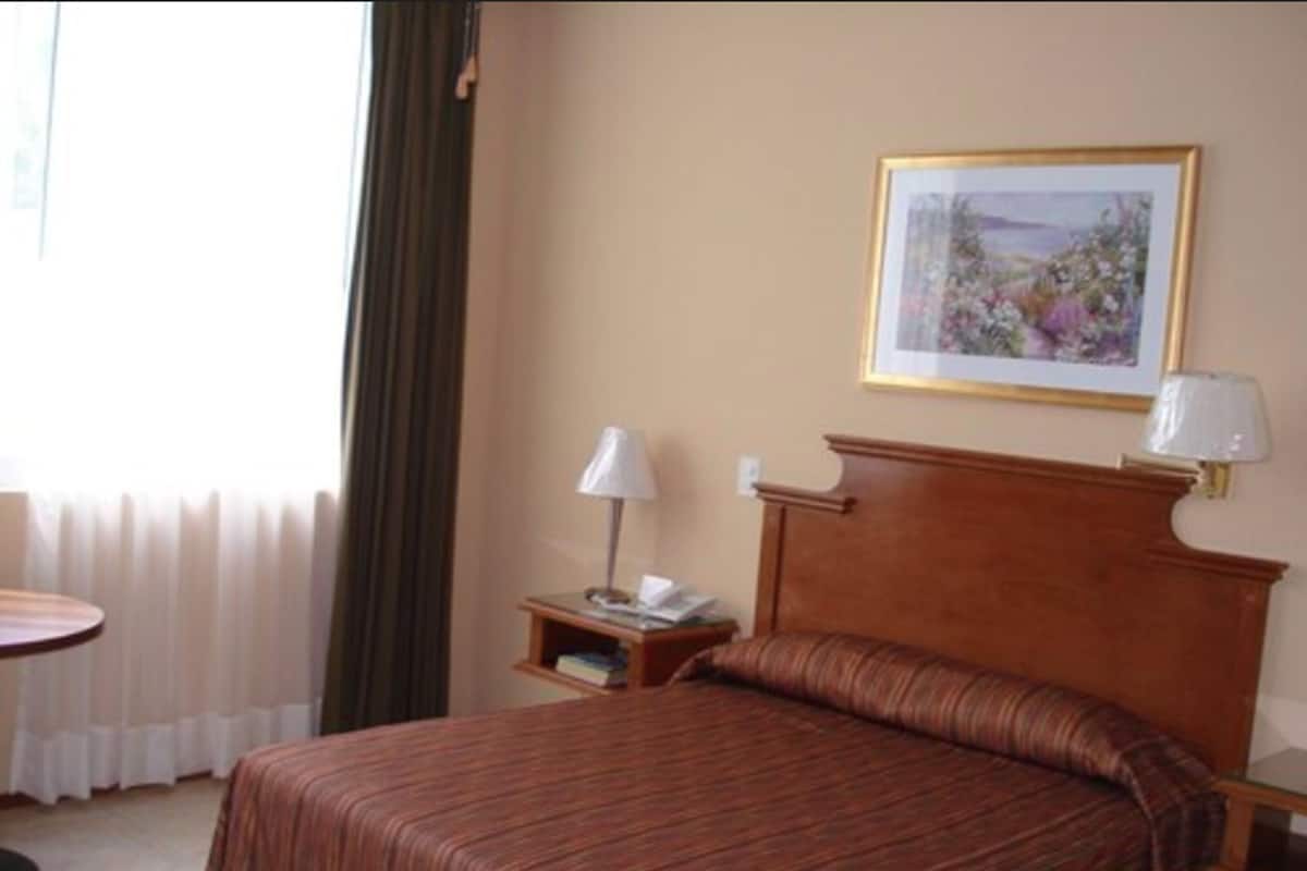 A hotel room with a bed, a bedside table with a lamp, a framed picture on the wall, and a window with curtains.