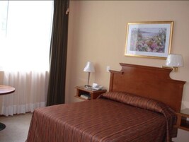 Habitación de hotel con cama, mesita de noche con lámpara, cuadro enmarcado en la pared y ventana con cortinas.