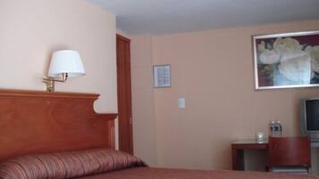 Una habitación de hotel con cama, lámpara, un cuadro, televisor y una mesita.