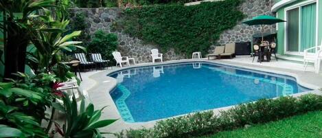 A rectangular swimming pool with a stone wall and greenery around it. There are lounge chairs and a table with umbrellas.