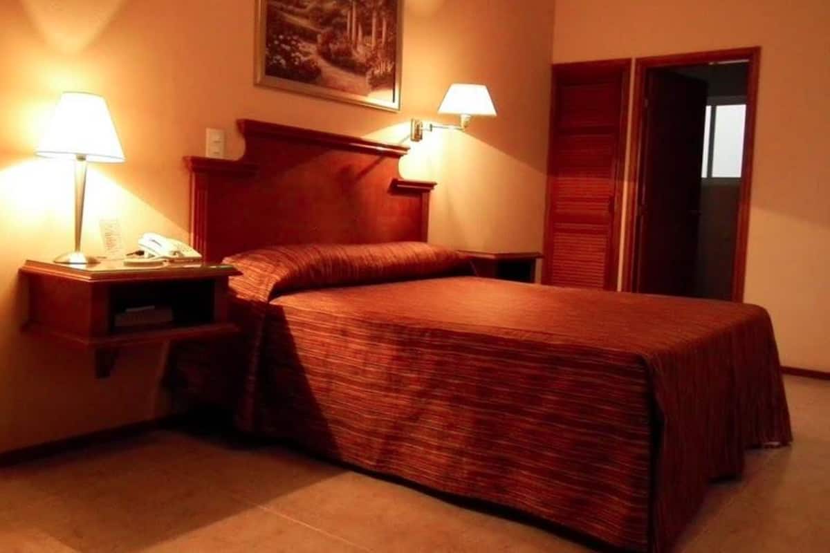 A hotel room with a bed, two bedside tables, a nightstand, a lamp, and a painting on the wall.