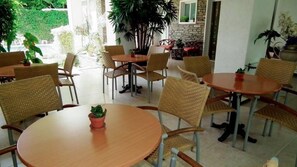 Zona pranzo con tavoli rotondi e sedie in vimini, circondata da piante in vaso e un muro di pietra.