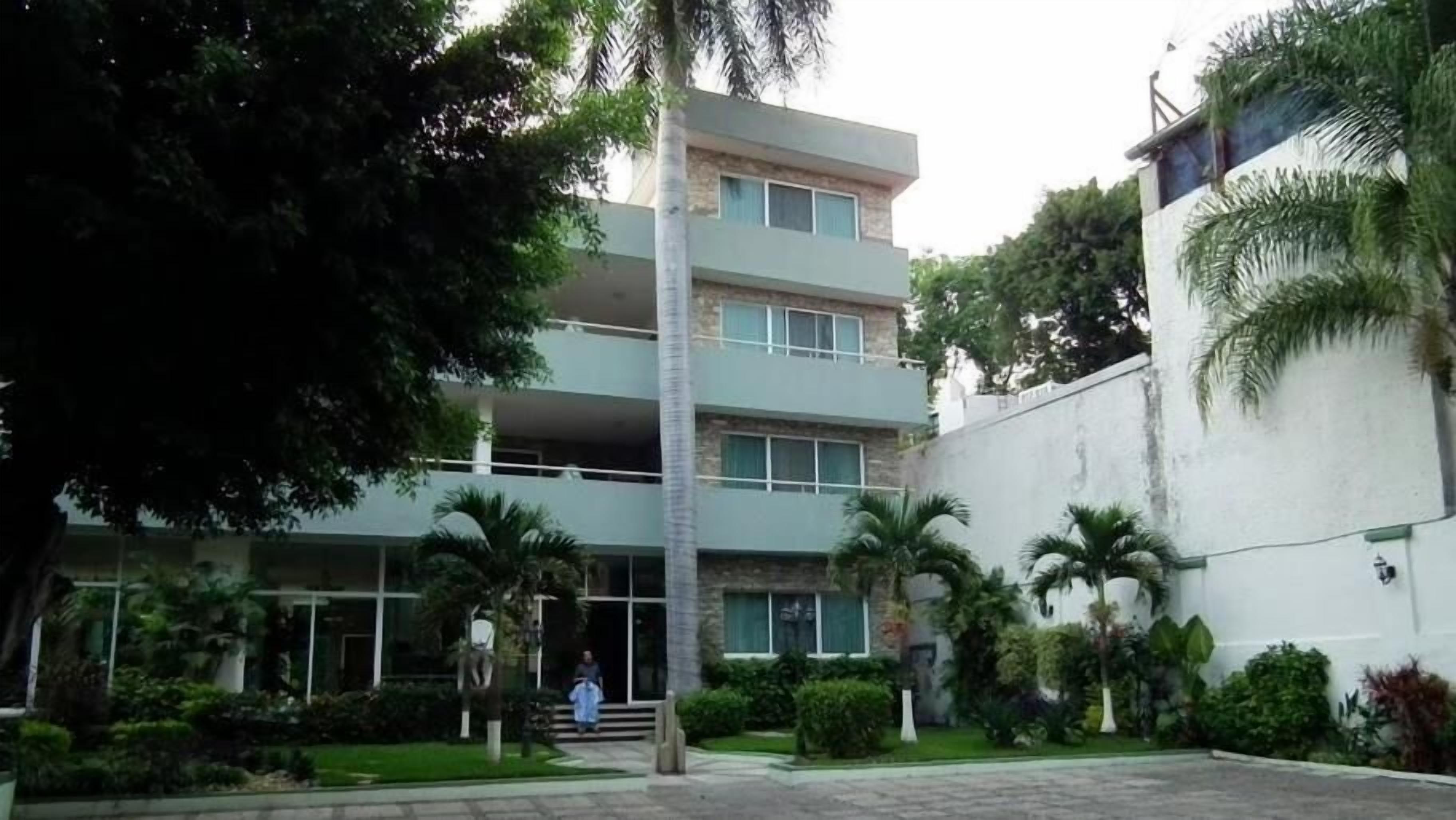 Un edificio de varios pisos con diseño moderno, que destaca una escalera central y un jardín bien cuidado con palmeras y arbustos.