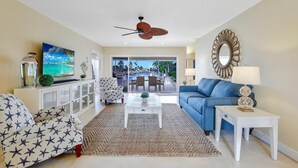 A spacious living room with a large TV, a ceiling fan, and a view of the outdoor pool area. - Waterfront Home With Pool and Foosball Table (Marco Island)