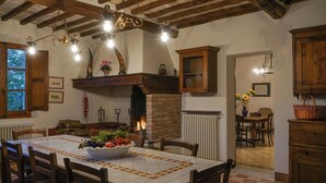 A dining area with a fireplace, wooden ceiling beams, and a table set with a bowl of fruit. - Villa La Pievina (Asciano)