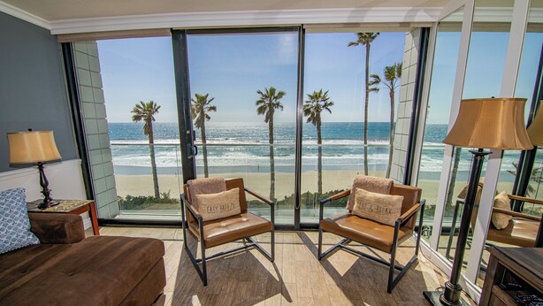 Een kamer met een groot raam met uitzicht op een strand, twee leren fauteuils met kussens, een bruine bank en een lamp.