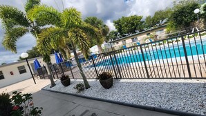 Una piscina con una valla negra, rodeada de guijarros blancos, palmeras y un edificio al fondo.