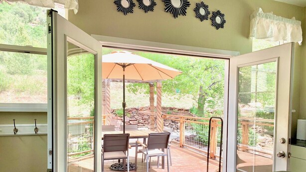 An open sliding glass door leading to an outdoor patio with a table and chairs, a wooden deck, and a stone wall. - The Fun House! Hot tub, Espresso Bar, Views, Foosball, Fire Pit, More! (Durango)