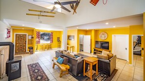 A spacious living area with a sofa, coffee table, and a fireplace. There is a ceiling fan, a TV, and a door leading to another room. - The Fun House! Hot tub, Espresso Bar, Views, Foosball, Fire Pit, More! (Durango)