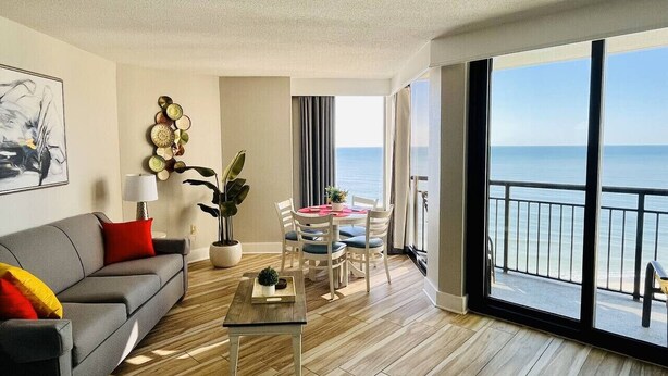 Ein modernes Wohnzimmer mit grauem Sofa, einem Couchtisch und einem Essbereich mit Stühlen und Tisch. Ein großes Fenster bietet Blick auf das Meer.