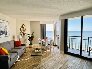 A modern living room with a grey sofa, a coffee table, and a dining area with chairs and a table. There is a large window with a view of the sea. - Absolute Beach. Oceanfront. Pet Friendly. (Myrtle Beach)