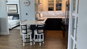 A modern kitchen with a dining area, a clock on the wall, and a view into the adjacent bedroom.