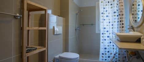 Een badkamer met toilet, douche en wastafel. Er is een houten schap en een douchegordijn met stippen.