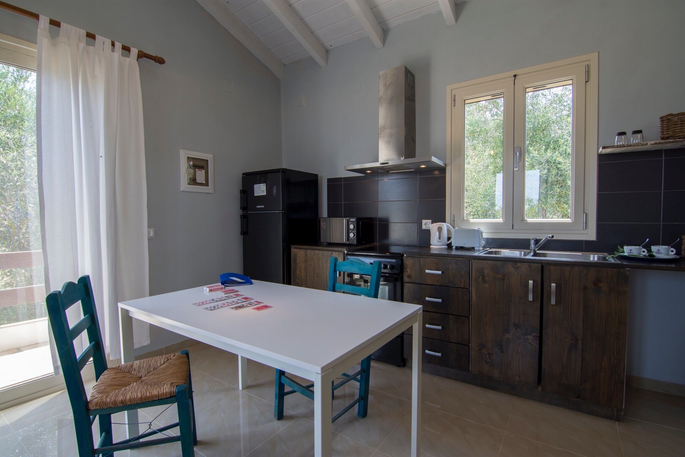 Uma cozinha com mesa e cadeiras brancas, geladeira preta e uma janela com cortinas.