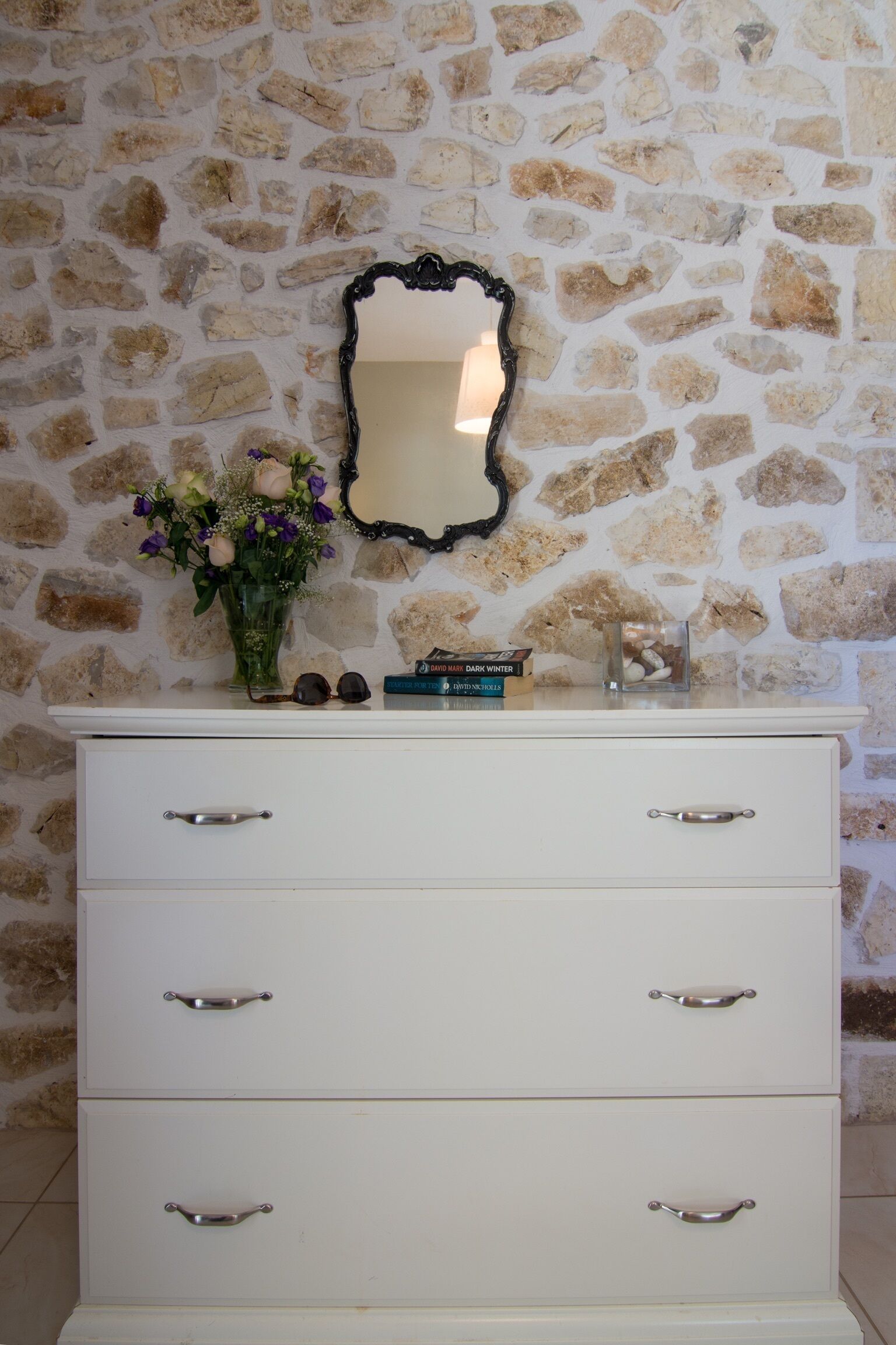 A white dresser with drawers, a mirror, and a vase with flowers.