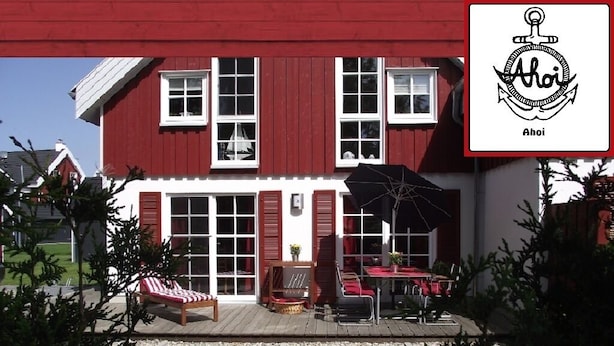 A red house with a deck, a lounge chair, and a table with an umbrella. - Holiday home in the heart of Greetsiel with whirlpool, sauna, fireplace and garden (Greetsiel)