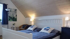 A bedroom with a sloped ceiling, a bed with blue bedding, two bedside lamps, a chair, a small table, and a potted plant. - Holiday home in the heart of Greetsiel with whirlpool, sauna, fireplace and garden (Greetsiel)