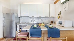 Una cocina compacta con armarios blancos, un refrigerador de acero inoxidable, una estufa y un microondas. Hay dos sillas con cojines rosados y una mesa con mantel azul.