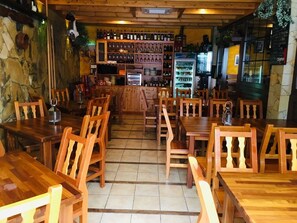 A restaurant with wooden tables and chairs, a bar area with shelves of bottles, and a refrigerator. - The House of Telegraphs (Vallehermoso)