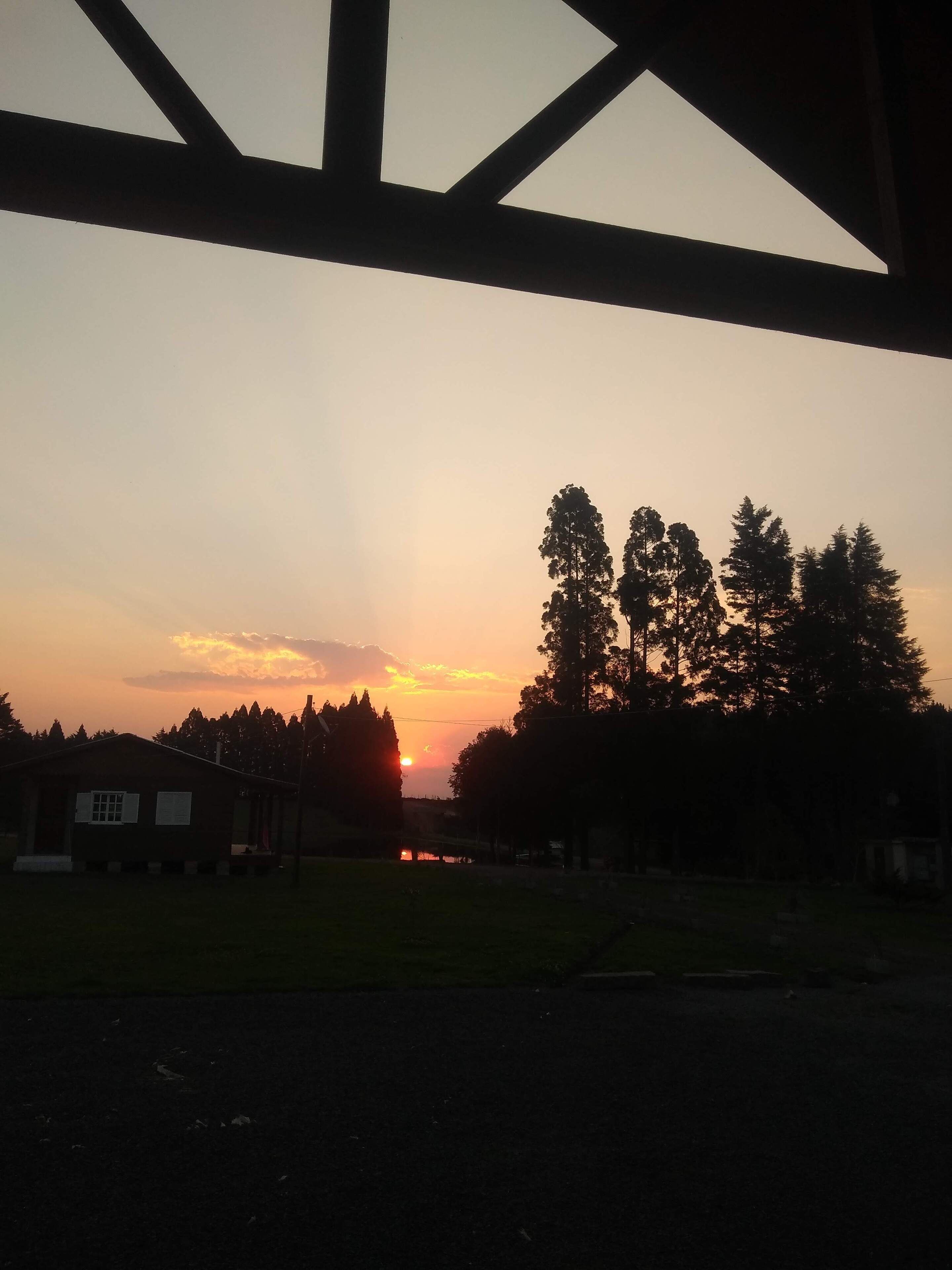 A sunset view from a covered area with a building and trees in the foreground.