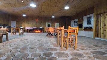 A spacious room with a wooden ceiling and stone flooring, featuring a large table and chairs, a fireplace, and a television.