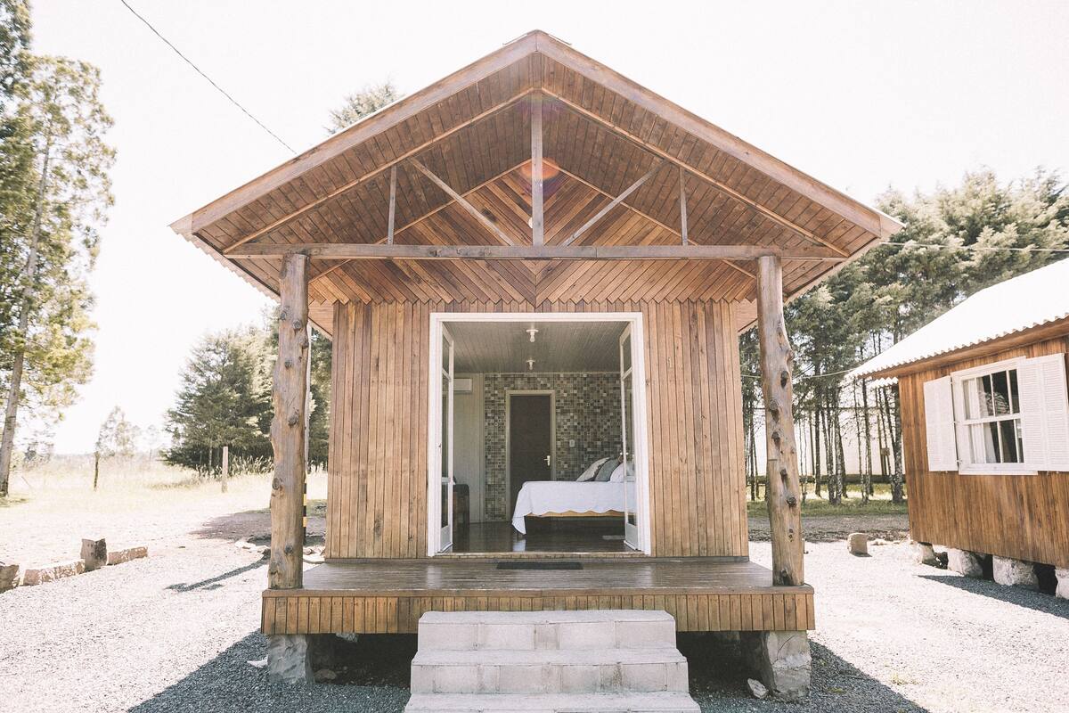 A wooden cabin with a porch, a bed inside, and a view of the surrounding trees.