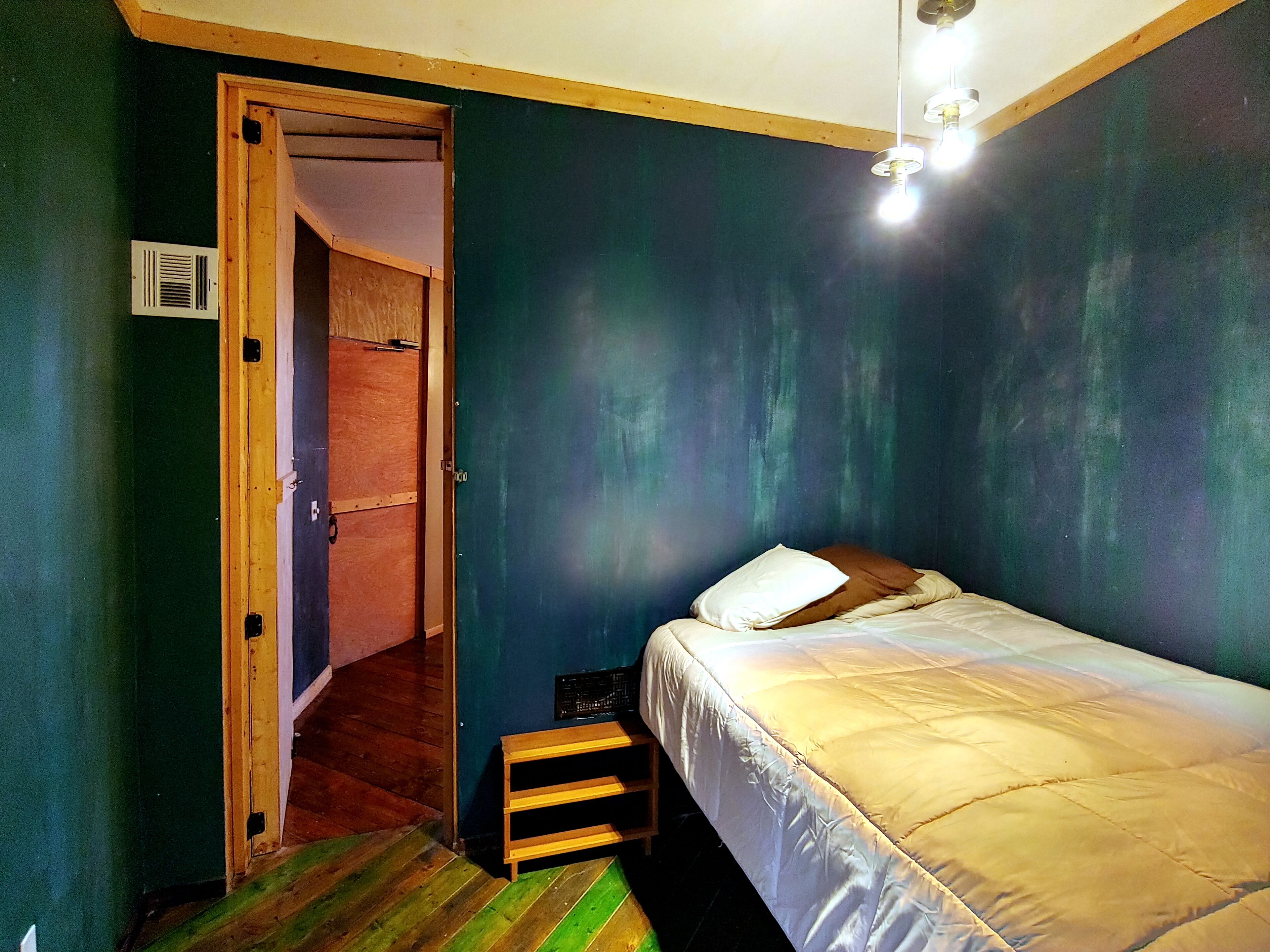 A small, narrow room with a single bed, a wooden door, and a small stool.