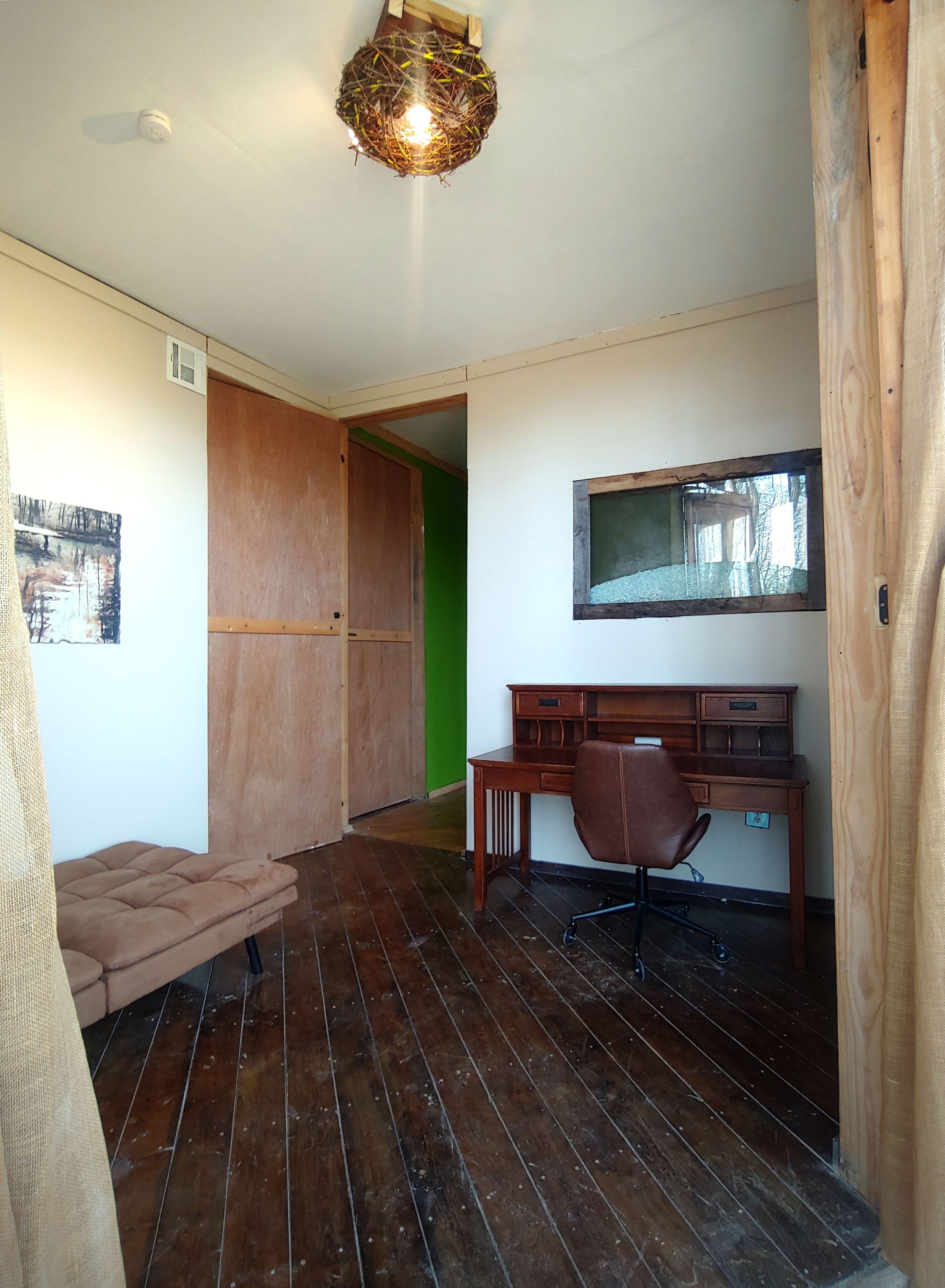 Una habitación con un escritorio de madera, una silla, un ventanal pequeño y una lámpara fijada a la pared.